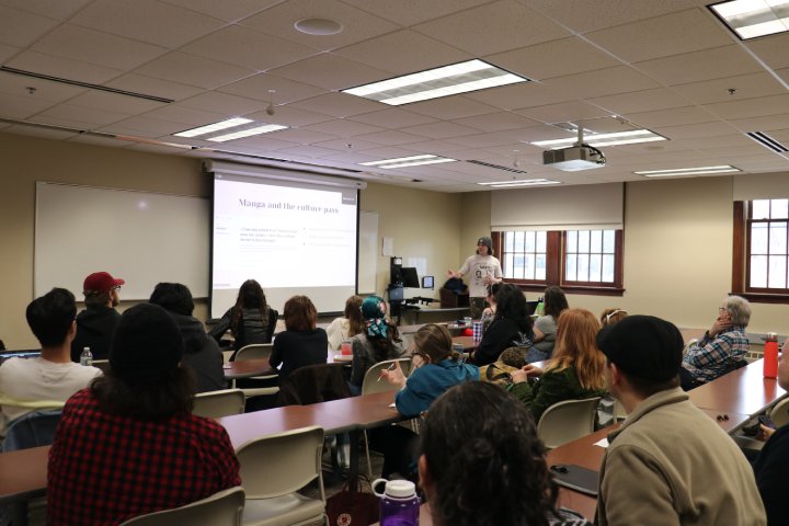 A presenter stands at the front of a classroom, giving a lecture on 'Manga and the Culture Pass' as projected on the screen. The audience is seated at long tables, attentively watching the presentation. The room is bright, with a mix of natural and artificial light, and the atmosphere feels informal and educational.
